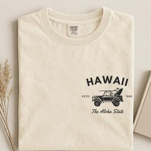 May include: Cream-colored t-shirt with a crew neck. The shirt features a black graphic of a vintage vehicle with a surfboard, the word "HAWAII", and the text "The Aloha State". The shirt also has the text "ESTD 1959".