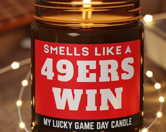 Puede incluir: Una vela encendida en un tarro de cristal marrón con una etiqueta roja que dice "Smells Like a 49ers Win" en texto blanco. La vela está sobre un pequeño posavasos de madera, con una guitarra y un libro en el fondo. El texto "My Lucky Game Day Candle" está en la parte inferior.