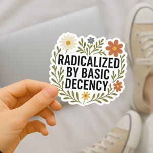 May include: A white sticker with the text "RADICALIZED BY BASIC DECENCY" in a floral wreath. The sticker is held in a hand, with a laptop and beige trainers in the background.