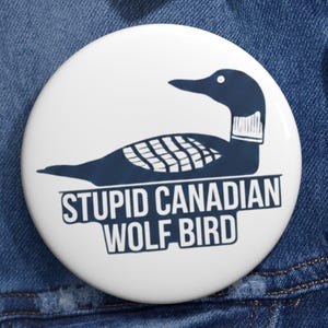 May include: A white button with a navy blue illustration of a loon and the text "STUPID CANADIAN WOLF BIRD" in black. The button is pinned to a denim jacket.