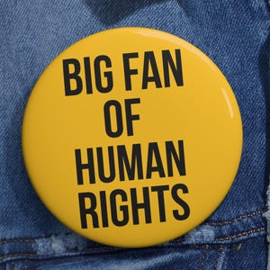 May include: A yellow button with the text "BIG FAN OF HUMAN RIGHTS" in black lettering. The button is pinned to a blue denim jacket. The button is round and has a glossy finish.