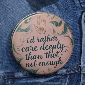 May include: A circular pinback button featuring a pink and teal marbled background with a line-art illustration of a hand with an eye in the palm. The text reads, I'd rather care deeply than not enough, written in a dark green cursive script font.
