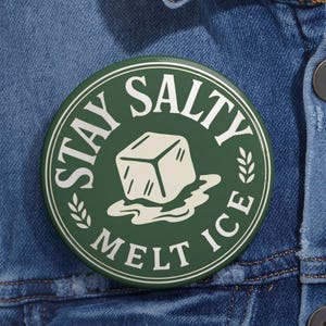 May include: A circular pinback button with a dark green background and white text that reads STAY SALTY MELT ICE. The center features a graphic of a white ice cube melting into a puddle, flanked by two small white leaf sprigs within a circular border.