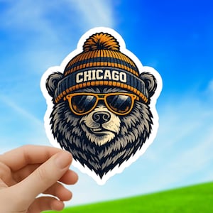 May include: A sticker depicting a cartoon bear wearing a navy blue and orange striped beanie with the word "CHICAGO" and orange sunglasses. The bear has a smiling expression. The background is a blue sky and green field.