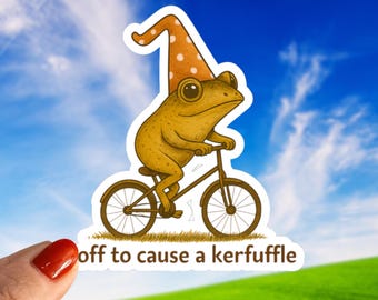 May include: A sticker featuring a whimsical illustration of a frog riding a bicycle, wearing a pointed hat with white polka dots. The text "off to cause a kerfuffle" is below the frog. The background shows a blue sky and green grass.