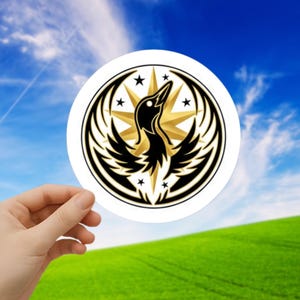 May include: A round white sticker featuring a black bird design with gold accents and stars. The bird is centered within a black and gold circular frame. The background shows a blue sky and green field.
