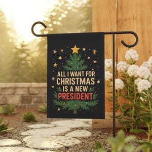 May include: Black garden flag with the text "ALL I WANT FOR CHRISTMAS IS A NEW PRESIDENT" in white, green, and red. The design includes a Christmas tree and stars. The flag is displayed on a black metal stand in a garden setting.