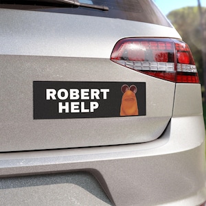 May include: A black rectangular bumper sticker with the words "ROBERT HELP" in white block letters. A cartoon character with brown ears and a tan face is on the right side of the sticker. The sticker is attached to the rear of a silver car.