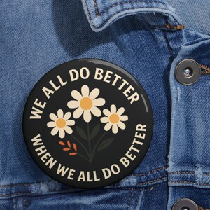 May include: A round black button with the text "WE ALL DO BETTER WHEN WE ALL DO BETTER" in white. The button features a floral design with three white daisies with yellow centers and green stems.