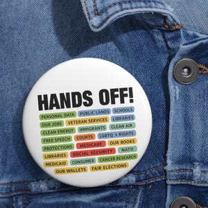 Puede incluir: Un botón blanco con las palabras "HANDS OFF!" en negrita negra. Debajo, varios rectángulos de colores con frases como "PERSONAL DATA", "OUR JOBS" y "FREE SPEECH".