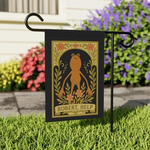 Op de afbeelding: Zwarte tuinvaandel met een bruine kikkerillustratie en de tekst "ROBERT, HELP". De kikker is omlijst door gouden en oranje bloemen- en bladeren ontwerpen. De vlag wordt getoond op een zwarte metalen standaard in een tuin.