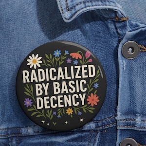 Może przedstawiać: Czarny guzik z napisem "RADICALIZED BY BASIC DECENCY" w kolorze białym i zielonym, otoczony kolorowymi kwiatami i liśćmi. Guzik jest umieszczony na dżinsowej kurtce.