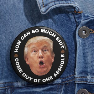 Könnte beinhalten: Ein schwarzer Button mit dem Gesicht eines Mannes mit blonden Haaren und dem Text "HOW CAN SO MUCH SHIT COME OUT OF ONE ASSHOLE" in Weiß. Der Button befindet sich auf einer blauen Jeansjacke.