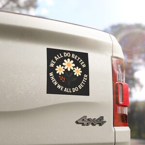 Pode incluir: Um autocolante quadrado preto com o texto "WE ALL DO BETTER WHEN WE ALL DO BETTER" a rodear um desenho floral de três margaridas. O autocolante está afixado na parte traseira de um veículo branco.