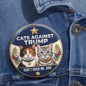 May include: A round pinback button featuring a dark blue and cream background with a gold star at the top. It displays two cats, one calico and one silver tabby, surrounded by floral accents. The text reads CATS AGAINST TRUMP and DON'T GRAB ME, BRO.