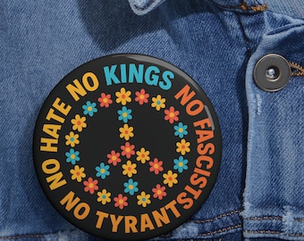 Könnte beinhalten: Ein runder, schwarzer Button mit dem Text "NO HATE NO KINGS NO FASCISTS NO TYRANTS" in Gelb, Blau und Orange. Ein Friedenszeichen aus bunten Blumen befindet sich in der Mitte. Der Button ist an einer Jeansjacke befestigt.