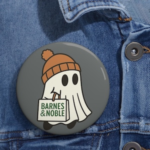 May include: A round button with a cartoon ghost wearing an orange beanie and carrying a shopping bag. The bag has the text "BARNES & NOBLE" in green and white. The button is set against a gray background and a blue denim jacket.