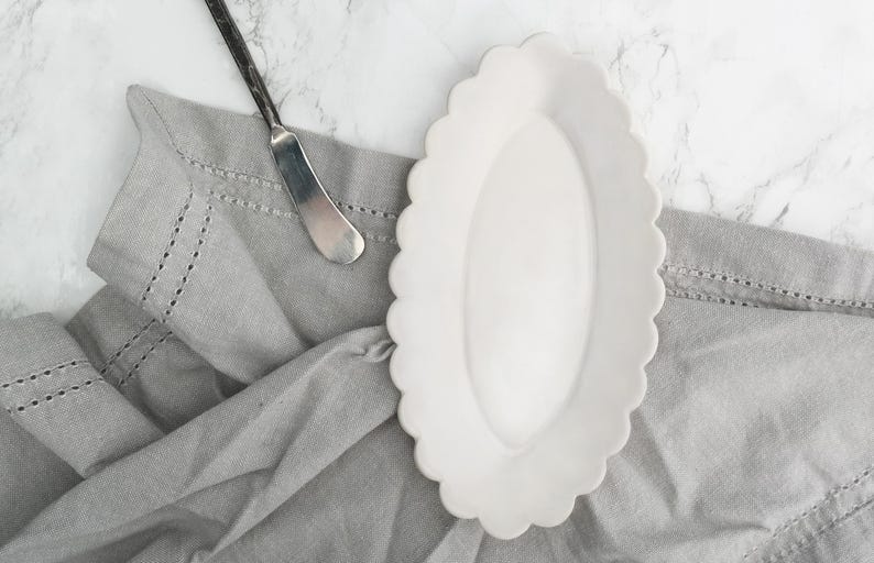 K&ouml;nnte beinhalten: Eine wei&szlig;e Keramik-Butterdose mit gewelltem Rand. Die Dose ist oval geformt und hat eine glatte, gl&auml;nzende Oberfl&auml;che.