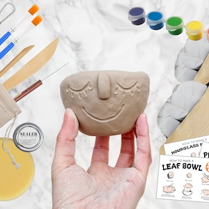 May include: A DIY pottery kit featuring a clay bowl with a smiling face design, paint pots, tools, clay, and instructions. The kit includes brushes, a sponge, and a sealer. The bowl is held in a hand.