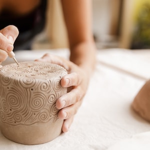 May include: A person carving a decorative pattern into a cylindrical, light brown ceramic piece. The design features swirling, spiral motifs. A second, unfinished ceramic piece is visible in the background.