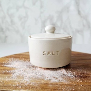 May include: A cream-colored ceramic salt cellar with a lid, labeled "SALT" in embossed letters. The salt cellar sits on a wooden cutting board, with a scattering of pink salt granules. The background is a white marble pattern.
