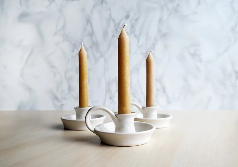 May include: Three white ceramic candle holders with handles, each holding a tall, slender, beige candle.