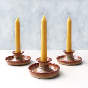 May include: Three yellow candles are displayed on brown ceramic candle holders. The candles are tall and slender, with a white wick at the top. The candle holders have a simple design with a round base and a raised center.