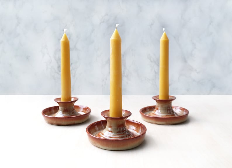 May include: Three yellow candles are displayed on brown ceramic candle holders. The candles are tall and slender, with a white wick at the top. The candle holders have a simple design with a round base and a raised center.