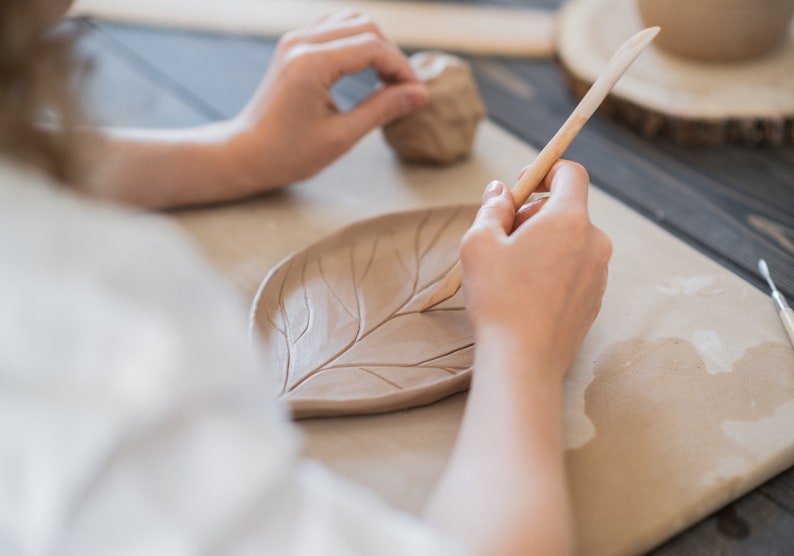 May include: A person's hands are shown working with clay, shaping a leaf-shaped dish with a wooden tool. The clay is a light brown color, and the dish features detailed leaf vein impressions. A small ball of clay sits nearby.