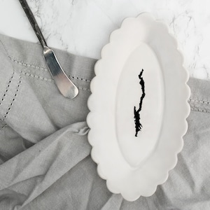 May include: A white oval-shaped plate with a scalloped edge, featuring a black silhouette of a lake or island. The plate is on a gray linen napkin.