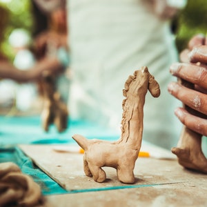 May include: A light brown clay giraffe sculpture stands on a wooden board, with a second giraffe sculpture being worked on in the background. The giraffe has a long neck and small legs. The scene suggests a pottery class or art project.