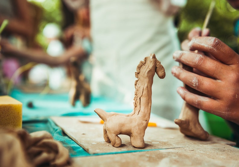 May include: A light brown clay giraffe sculpture stands on a wooden board, with a second giraffe sculpture being worked on in the background. The giraffe has a long neck and small legs. The scene suggests a pottery class or art project.