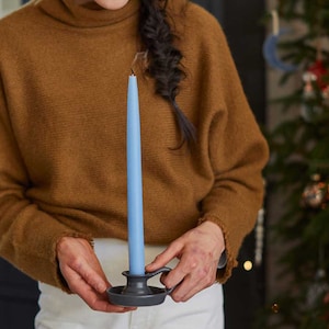 May include: A person holding a blue candle in a black candlestick. The candle is lit and the person is wearing a brown sweater.