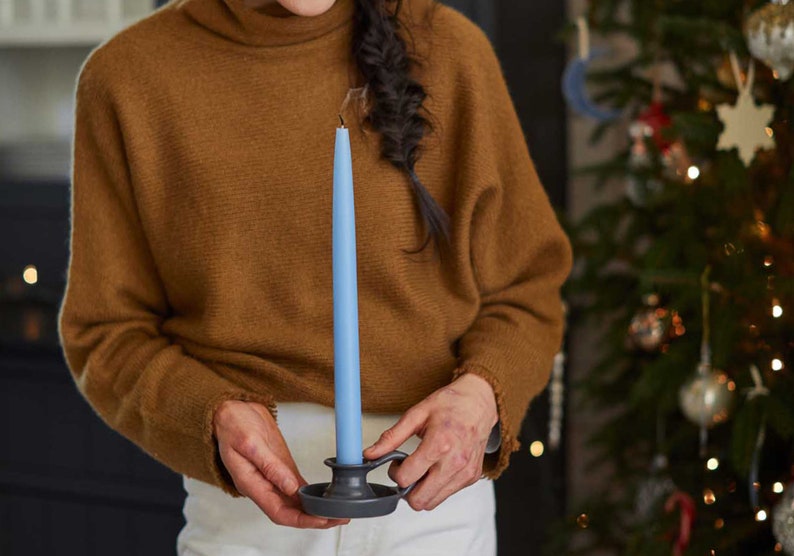 May include: A person holding a blue candle in a black candlestick. The candle is lit and the person is wearing a brown sweater.