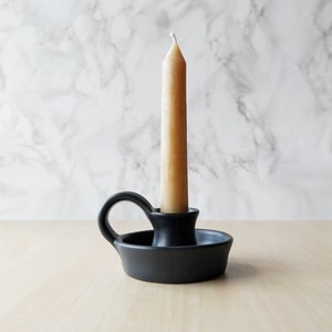 May include: A black ceramic candle holder with a single candle. The candle is a light brown color and is sitting in the holder.