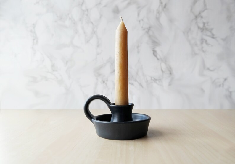 May include: A black ceramic candle holder with a single candle. The candle is a light brown color and is sitting in the holder.