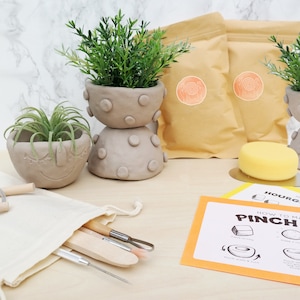 May include: A pottery kit with clay pots, tools, and instructions. The kit includes two stacked gray clay pots with green plants, a smiling clay pot, tools in a canvas bag, and instructions for making a pinch pot.