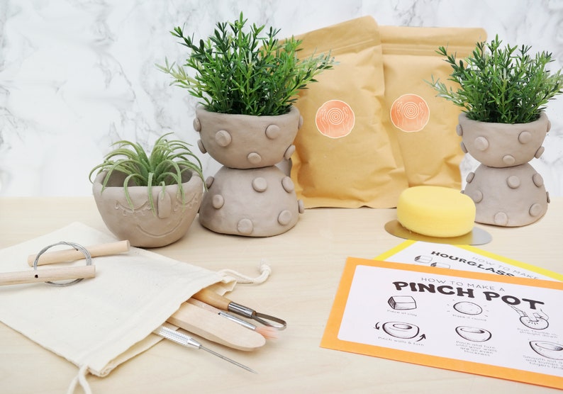 May include: A pottery kit with clay pots, tools, and instructions. The kit includes two stacked gray clay pots with green plants, a smiling clay pot, tools in a canvas bag, and instructions for making a pinch pot.