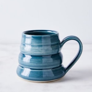Op de afbeelding: Een teal keramische mok met een uniek, geribbeld ontwerp. De mok heeft een grote, gebogen handgreep en een glanzende afwerking. De handgemaakte mok is perfect voor koffie, thee of een warme drank. De mok is ongeveer 10 cm hoog.