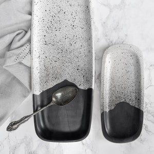 May include: Two rectangular serving trays with a speckled white and black design. The bottom half of each tray is solid black. A silver spoon rests on the larger tray, and a gray cloth is draped nearby. The trays are on a marble surface.