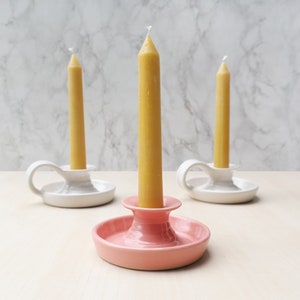 May include: Three ceramic candle holders with a single yellow candle in each. The holders are white with a handle and a pink holder with a wider base.
