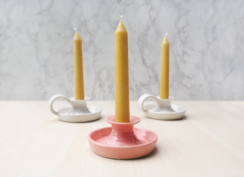 May include: Three ceramic candle holders with a single yellow candle in each. The holders are white with a handle and a pink holder with a wider base.
