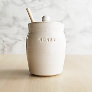 May include: Cream-colored ceramic honey pot with a lid and a wooden honey dipper. The word "HONEY" is embossed on the front. The pot sits on a light-colored wooden surface against a marble background.