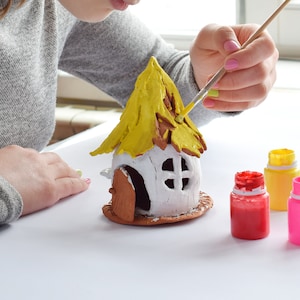 May include: A person painting a small, white ceramic house with a yellow roof. The house has a brown door and window. Several small bottles of paint in red, pink, yellow, green, and orange are visible.
