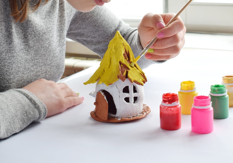 May include: A person painting a small, white ceramic house with a yellow roof. The house has a brown door and window. Several small bottles of paint in red, pink, yellow, green, and orange are visible.