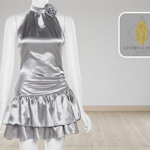 May include: A silver satin dress on a mannequin. The halter-style top features a keyhole detail and a decorative rose. The skirt has a layered, ruffled hem. The dress is displayed in a studio setting with a logo in the background.