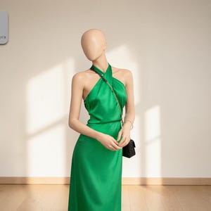 May include: A green halter dress with a flowing skirt. The dress is made of a shiny fabric and is worn by a mannequin. A small black purse hangs from the mannequin's shoulder. The dress has a simple, elegant design.