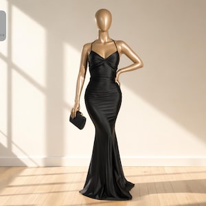 May include: A black, floor-length evening gown with a halter neckline and a fitted silhouette. The dress is displayed on a mannequin holding a small black clutch. The mannequin is gold-colored and stands on a wooden floor.