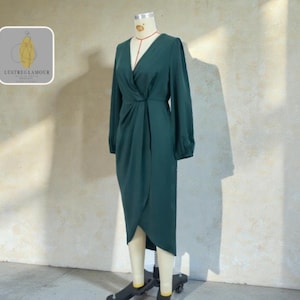 May include: A dark teal dress with long sleeves and a V-neck, displayed on a mannequin. The dress features a wrap-style design at the waist and a tulip hem. The mannequin is on a wheeled stand. The logo "Lustreglamour" is in the upper left corner.