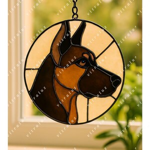 Puede incluir: Adorno de ventana de vidriera circular con el perfil de un pastor alemán. El diseño utiliza tonos marrones, beige y negro, con una cadena de metal negro. El texto "vitraluz" es visible. Ideal para colgar.